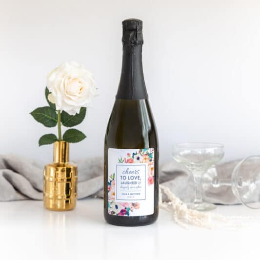 sticker on bubbly wine bottle with flower in vase and wine glasses in background